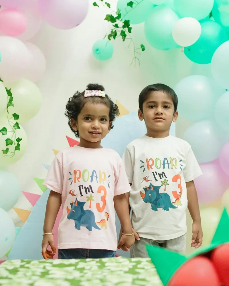 Two small children in matching dinosaur T-shirts in front of balloons